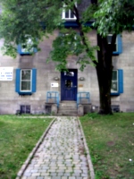 [picture: Cobbled path through grass past tree to blue door in grey stone building.]